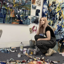 Laura Holmes squats in her studio, surrounded by abstract paintings on the walls, and tools and paints on the floor